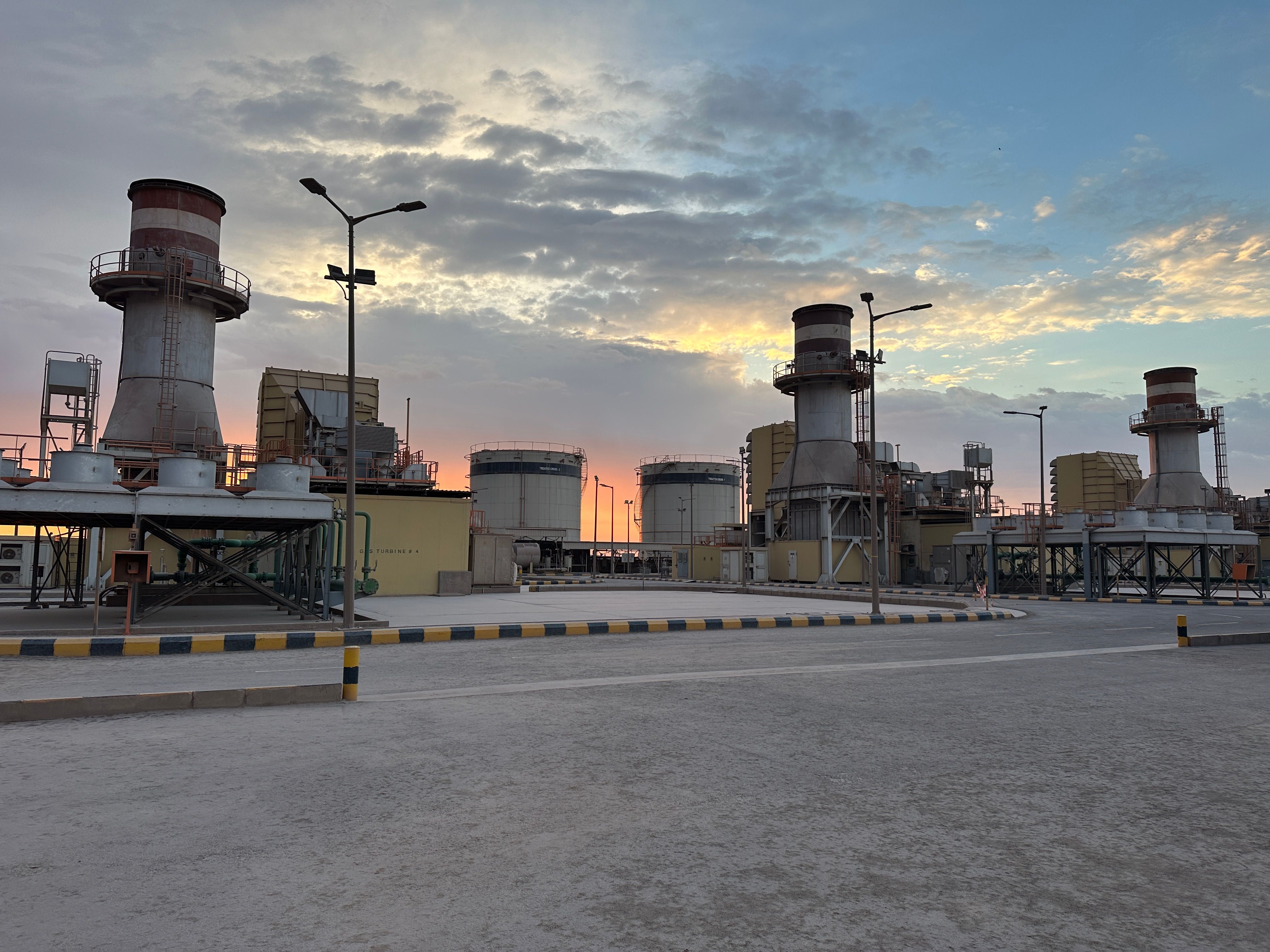 A power plant site at dusk, with turbine exhaust stacks and storage tanks against a partly cloudy sky.